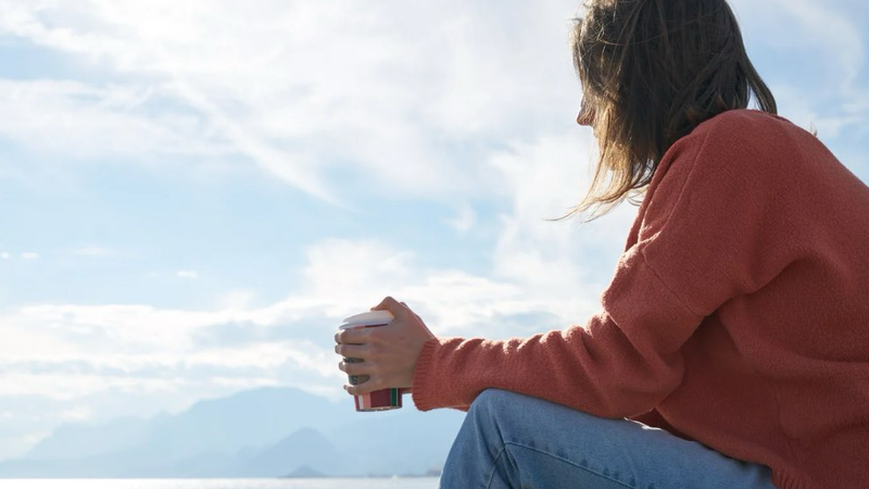 Person sitting quietly outdoors holding a warm drink, looking out over water and mountains in a moment of reflection.